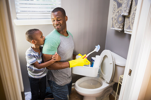 how to clean a porta potty | An African American man doing housework.  He is kneeling in the bathroom at the toilet bowl, wearing rubber gloves, holding cleaning supplies.  His little boy is keeping him company, hugging his arm.